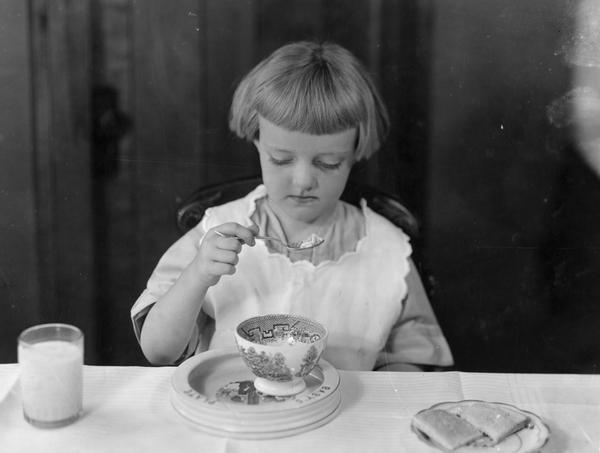 Young girl sitting at a table eating cereal(?) from a bowl. She also has a glass of milk and a plate with toast.