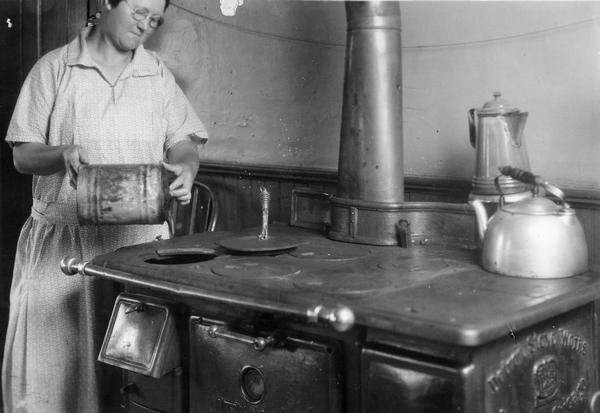 A woman pretends to pour kerosene on a hot stove as part of an International Harvester Agricultural Extension Department campaign to make families aware of farm and home hazards. The original caption reads: "Do not pour kerosene on a stove fire."