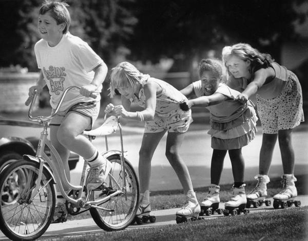 Kids make a "train," with the bicyclist in the front pulling three girls on roller skates along a sidewalk.