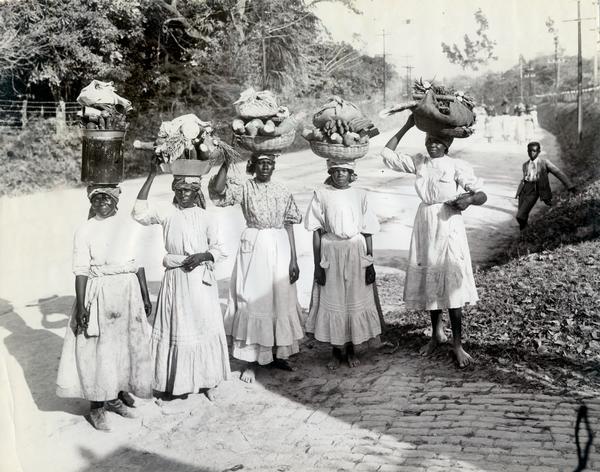 Group portrait of five Jamaican women on the road to market carrying baskets of goods on their heads.