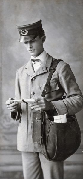 Carte-de-visite of a mailman dressed in his full uniform gazing at a piece of mail he is holding in his hands. He is carrying his mail pouch over his shoulder.
