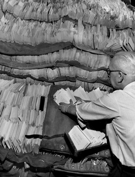 Man sorting envelopes in a mail room near tall stacks of mail.