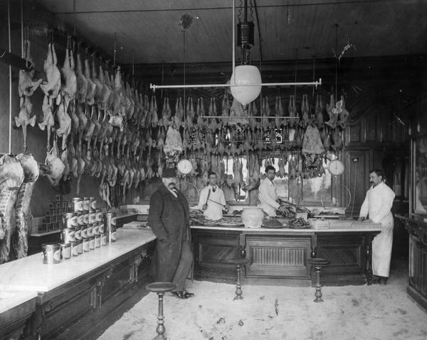 A customer waits while butchers stand behind the counter of a butcher shop, surrounded by meat hanging from the ceiling.