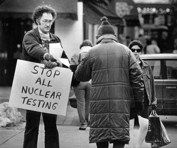 A man hands out information while holding a sign reading "Stop All Nuclear Testing" in front of the Reuss Federal Building.
