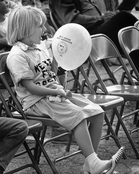 A young girl listens to a speech at the Racine Laborfest. She holds a balloon imprinted with the slogan, "Buy Union Buy American."