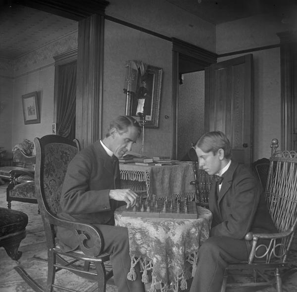 Reverend Henry Faville and his son, Ted, are seated around a small table playing chess in their home.