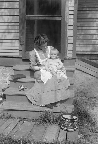 A mother sits with her baby on front steps in the sun surrounded by toys.