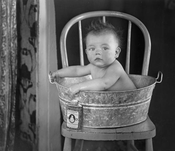 A baby sits in a galvanized tub holding on to the sides. The tub is on top of a chair with a can of talcum powder next to it.