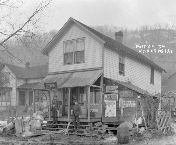Men, possibly postal workers, standing on the front steps of the local post office with crates and barrels in the foreground.