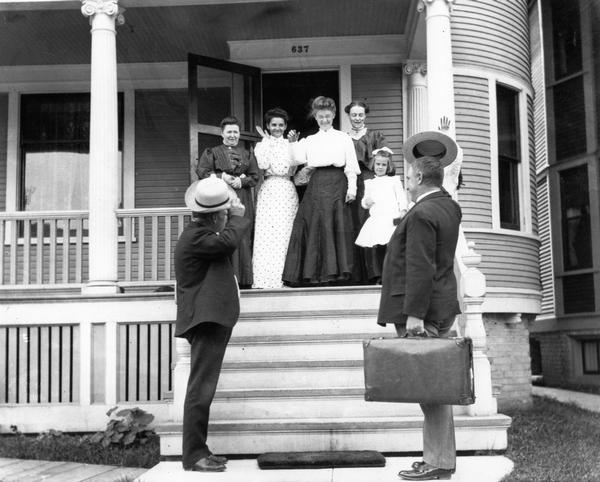 Mary E. (Mrs. Joseph) Smith, third from right, with family members on the porch, wave as two men at the bottom of the steps doff their hats.