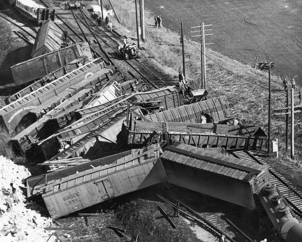 Elevated view of railroad cars laying across and around railroad tracks while workers look on and contemplate how to clean up the massive mess caused by the accident.