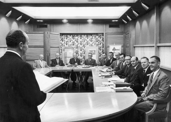 Men in suits are sitting around an oval-shaped table while another man with his back to the camera gives a presentation.