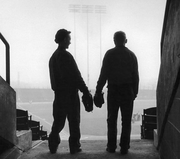 View from rear of two boys, in semi-silhouette, wearing baseball mitts standing and looking out over the baseball diamond from the seating area of County Stadium, home of the Milwaukee Braves.