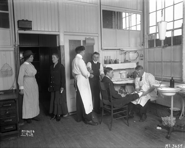 Doctors applying bandages to two men in a doctor's office or infirmary at International Harvester's McCormick Works. The men appear to be office workers. Two women are standing in the open doorway. One of the women has a bandage on her finger. The factory was owned by the McCormick Harvesting Machine Company until 1902.