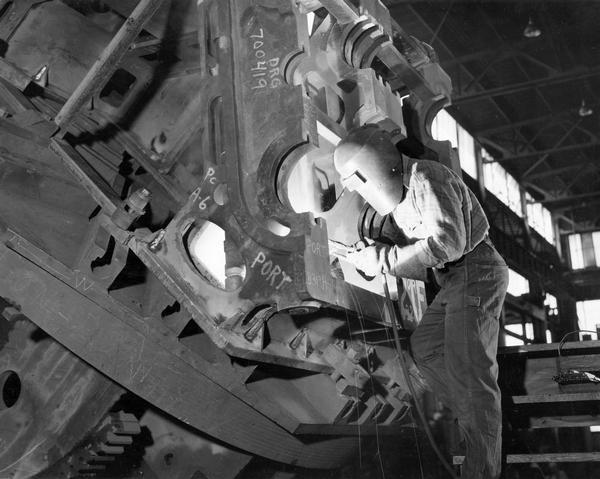 Man in protective gloves and masked helmet against heat and flame works on heavy machinery at the Falk Corporation, known for production of industrial gear drives.