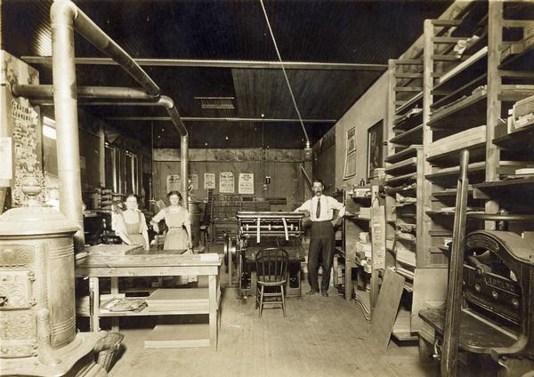 Interior view of the Brillion News Print Shop, where a printer and his assistants, two women, are posing around a press and work tables.