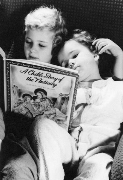 Two young children relax in their pajamas while reading "A Child's Story of the Nativity."
