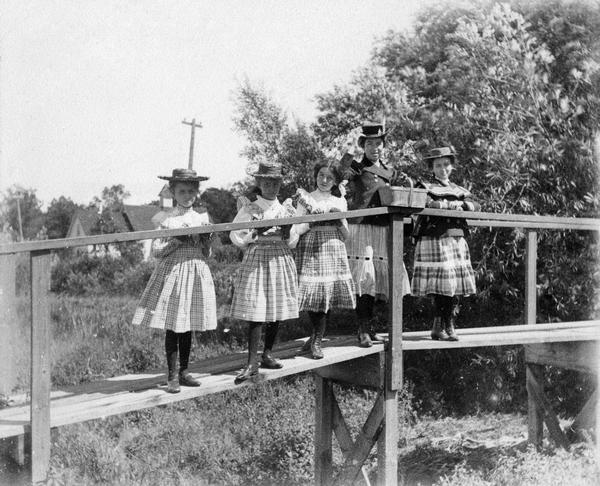 A young woman and four girls wearing frilly dresses and straw hats pose on a narrow wooden bridge.
