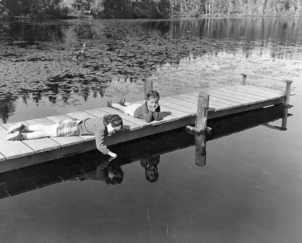 Two girls look into the water at their reflections as they lay sprawled on a wooden pier.