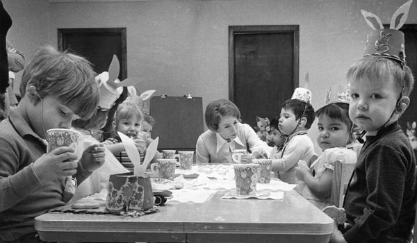 Party hats with rabbit ears are the headgear of choice at a childrens' party.  A woman is in deep conversation with one child while the others enjoy cookies and drinks.