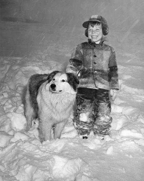 Winter scene with Mark Mueller, the photographer's son standing ankle deep in snow with a dog, and smiling as the snow continues to fall. The family was sledding north of Appleton near what is now Plamann Park.