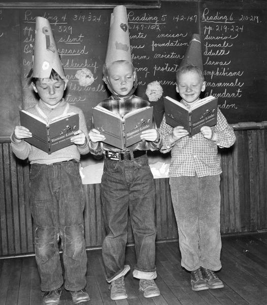 Three boys stand at the front of their classroom and read aloud the story, "The Funny Hats" while wearing funny hats.