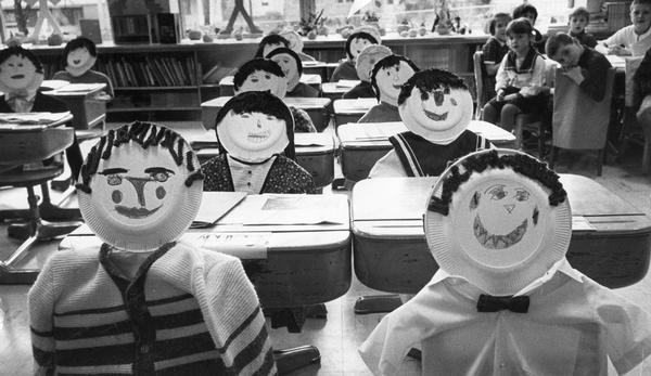 First grade students look at likenesses of themselves seated at their desks.  They created these by drawing their faces on paper plates, attaching them to their desks with coat hangers, and hanging clothes on the hangers.  This was done to surprise their parents at a school open house.