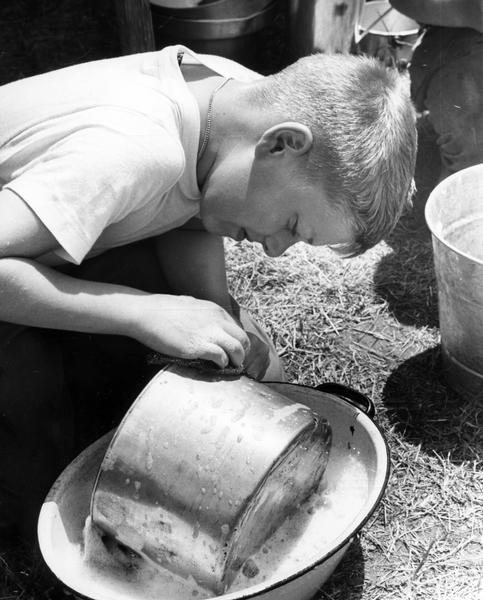 A boy kneeling down to scrub a pot that is resting in a basin of soapy water.