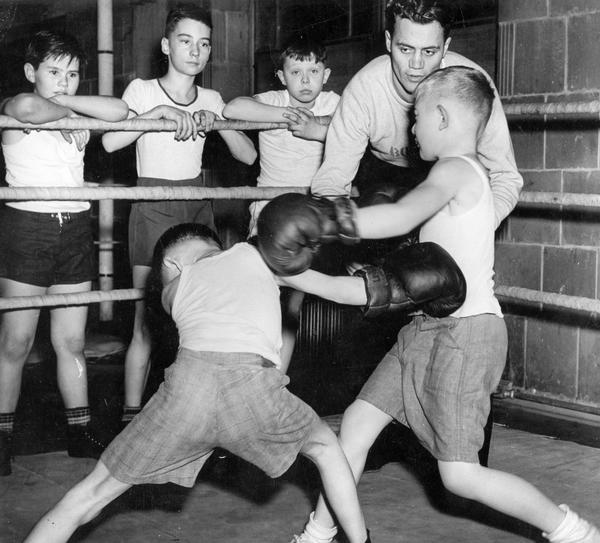 A man coaches boys in as they spar in a boxing ring with others watching closely.