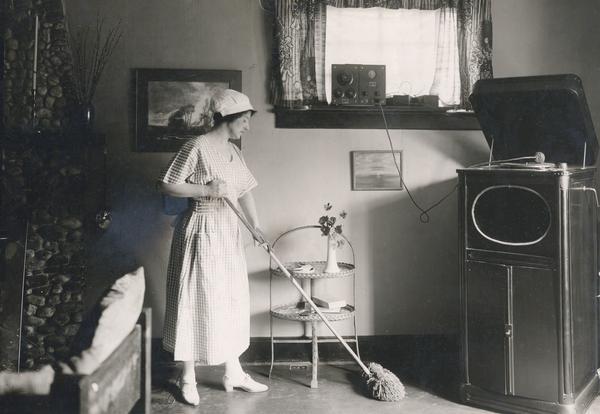 A woman dust mops the floor while listening to a radiophone that is perched on the window sill.