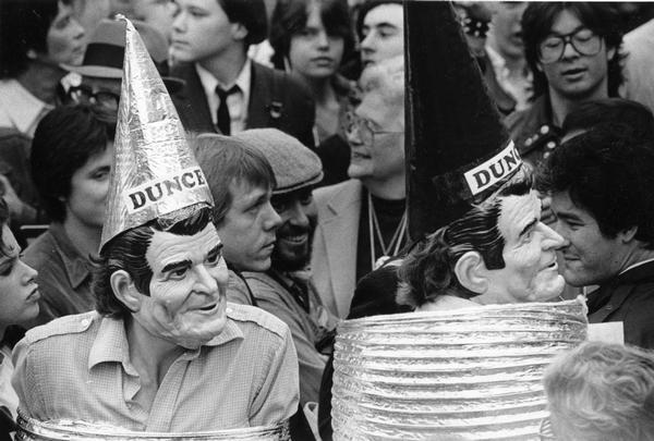 Men wearing Ronald Reagan masks and dunce caps make their way through a crowd.