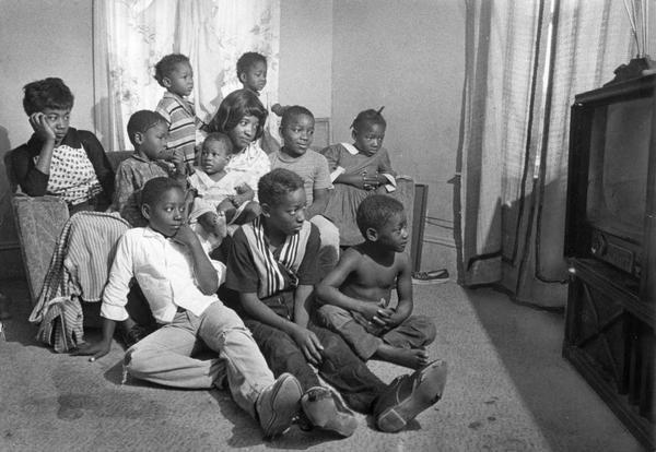 A woman sits with her ten children surrounding her and watches TV.