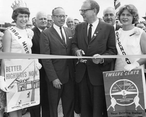 Governor John Reynolds, state highway officials, and beauty queens are in attendance as the ribbon is cut to open a new Wisconsin highway.  One of the young women holds a sign for National Highway Week that reads: "Better Highways Now Save Lives, Time, Money."