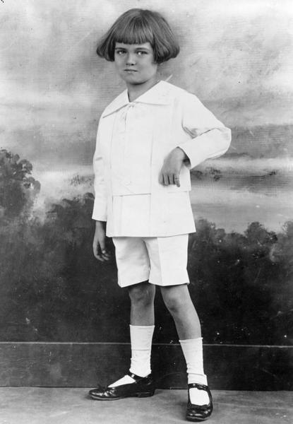 Full-length portrait in front of a painted backdrop of a young boy modeling a white linen suit with wide belt, embroidered collar, and silk cord tie. The boy has page or Dutch-boy bangs, with the hair at the side covering the ears.
