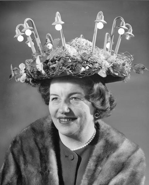 Mary Barnes, wearing a mink stole and pearls, modeling her outrageous hat. It is wired to light 10 street lights that are arranged around the brim.