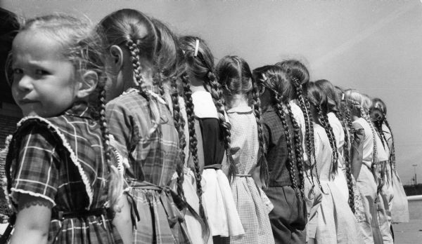 A line of girls, all with their hair in braids, stand with their backs to the camera.