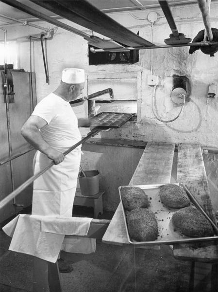 A baker, clad in apron, pulls a tray of freshly-baked rolls from the oven.