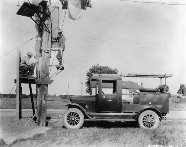 Telephone linemen working on scaffolds at different levels on a telephone pole as they install line in a rural area. Their International Harvester truck is parked in the grass nearby.