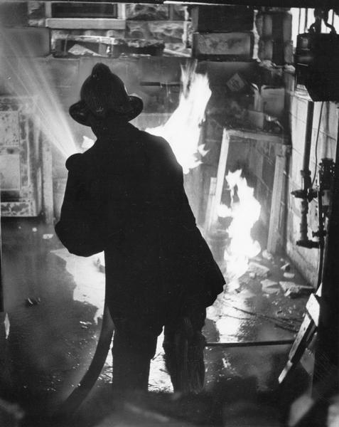 View towards a firefighter standing with his back to the camera as he is aiming a fire hose towards the flames.