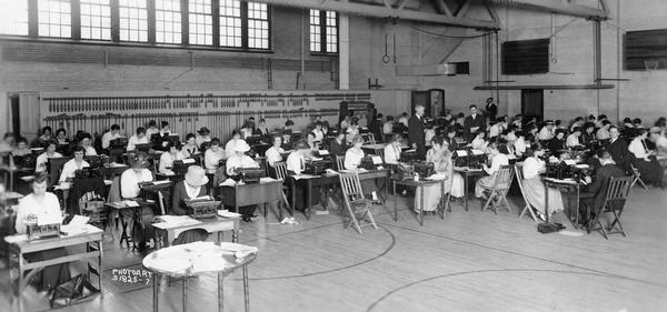 A large group of men and women are sitting at typewriters in a gymnasium to take the state civil service exam.