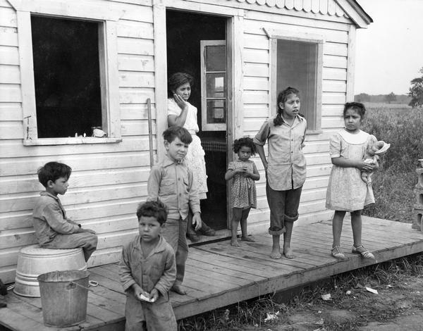 A Mexican woman and her six children are standing on the porch of the multiple-family housing provided to them by the pea cannery for which their husband and father works.