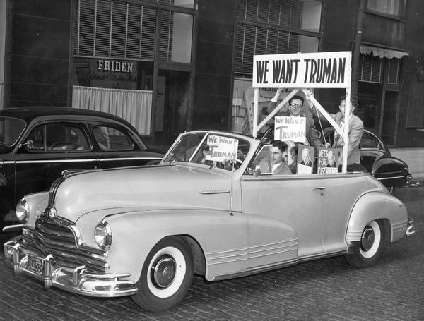 Men ride in a convertible with campaign signs proclaiming, "We Want Truman," and "Vets for Eisenhower."