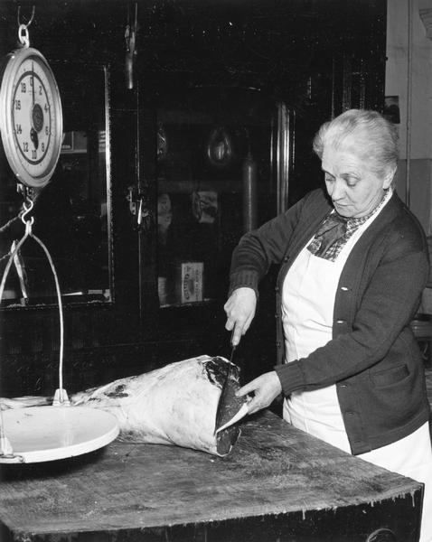 Next to a hanging scale, a large slab of meat on a butcher block is sliced into managable portions by apron-clad Mrs. Etta Parson Hocking.