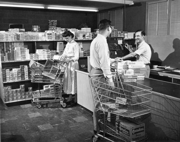 A cashier checking out a customer's purchases while another customer is filling her grocery cart with items at the D. Kurman company market.