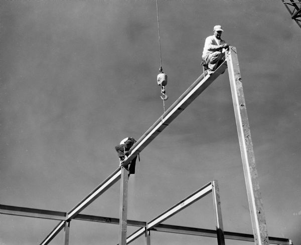 Construction workers sitting atop a steel girder placed by a crane, bolting it together at the corners.