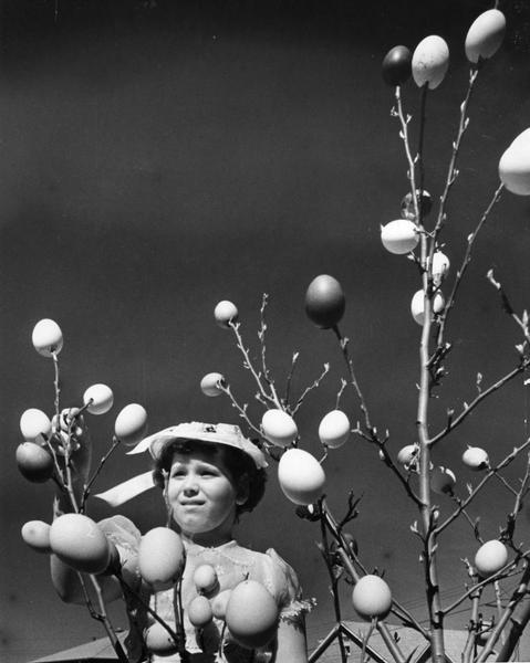 Bonnetted girl gazes at egg tree.