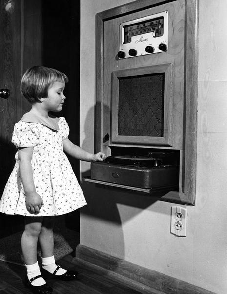 A little girl stands at the built-in home entertainment system consisting of a phonograph turntable, radio, and single speaker.