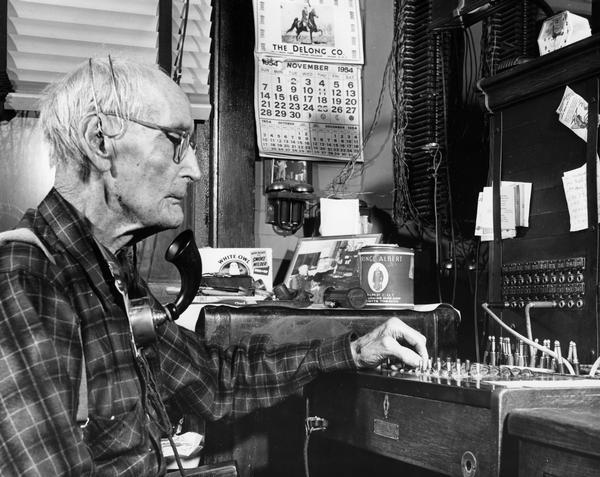 Henry Anderson runs a switchboard on his 90th birthday, with his smoking paraphernalia and a photograph of him at the switchboard on a table nearby.