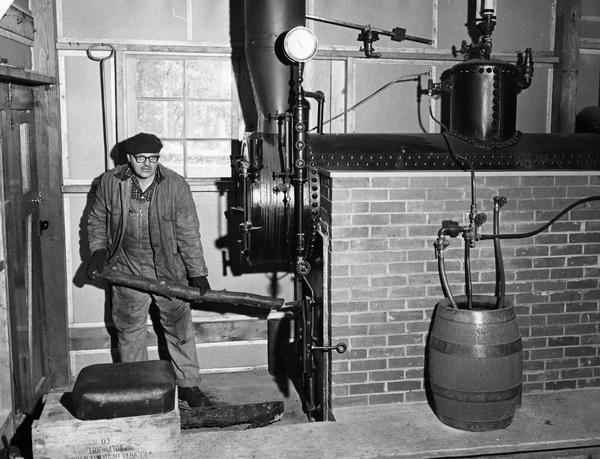 A man in work clothes gets ready to shove a log into the fire chamber of a boiler.