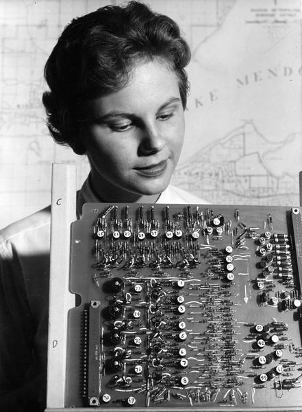 Jean Hoffman looks over a printed circuit board in the foreground, with a map of Madison, Wisconsin on the wall behind her.